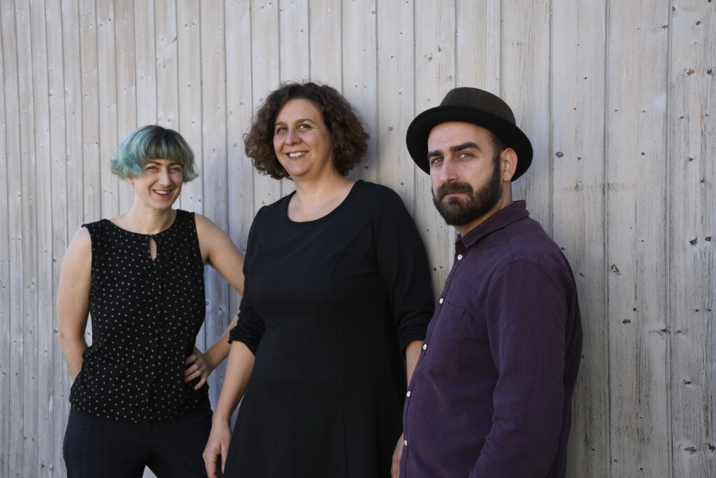 Gruppenfoto der Band: Angela, Elena und Larsen stehen vor einer Holzwand und lächeln in die Kamera